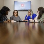 4 women sit at a conference table with a TV in the background