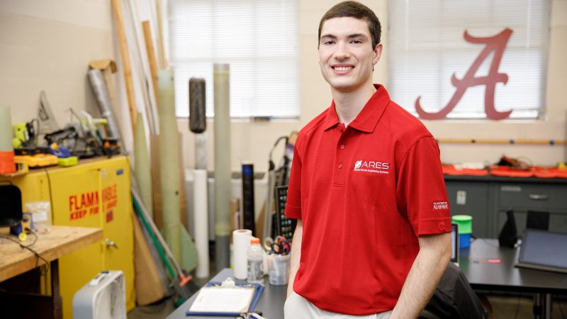 Peyton Strickland in a red shirt in a workshop with lots of rocketry items