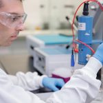 A man in a lab coat, eye gear, and gloves works with a machine