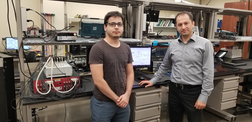 Two guys next to a counter and shelves filled with wires and a computer