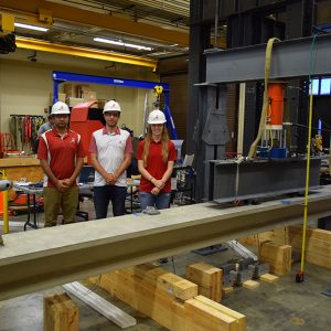 Three UA students stand in an on-campus lab with the cement beam in the foreground.