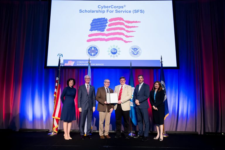 6 people stand on a stage with a booklet below a large screen