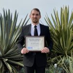 A man holds a plaque award in front of southwestern vegetation