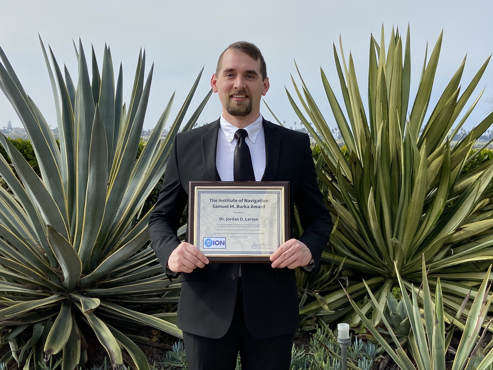 A man holds a plaque award in front of southwestern vegetation