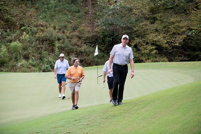4 men walking towards camera one in an orange shirt, golf flag in background