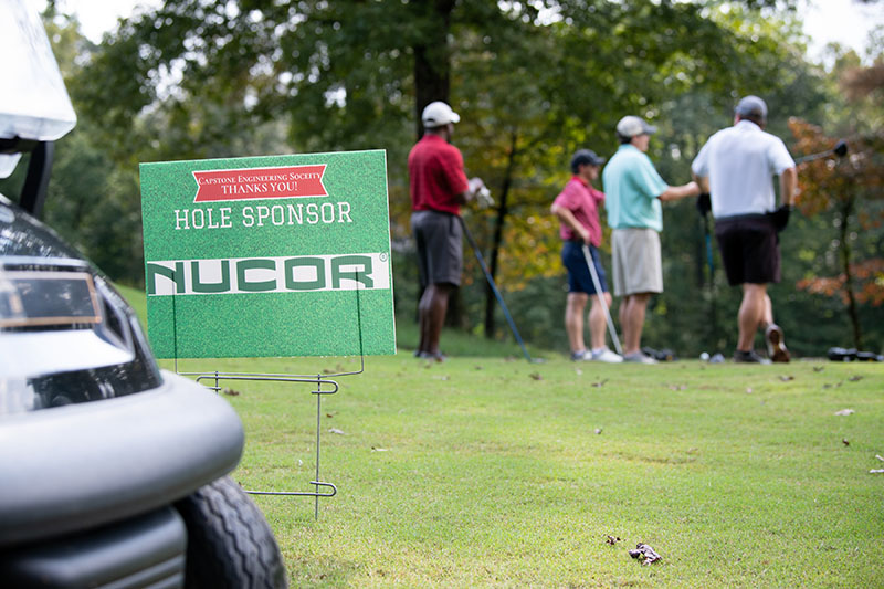 4 men in the distance with the camera focusing on a bit of golf cart and Nucor sign