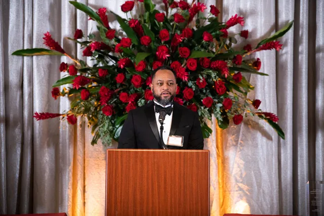 a man standing behind a podium with a huge bouquet of red flowers behind him