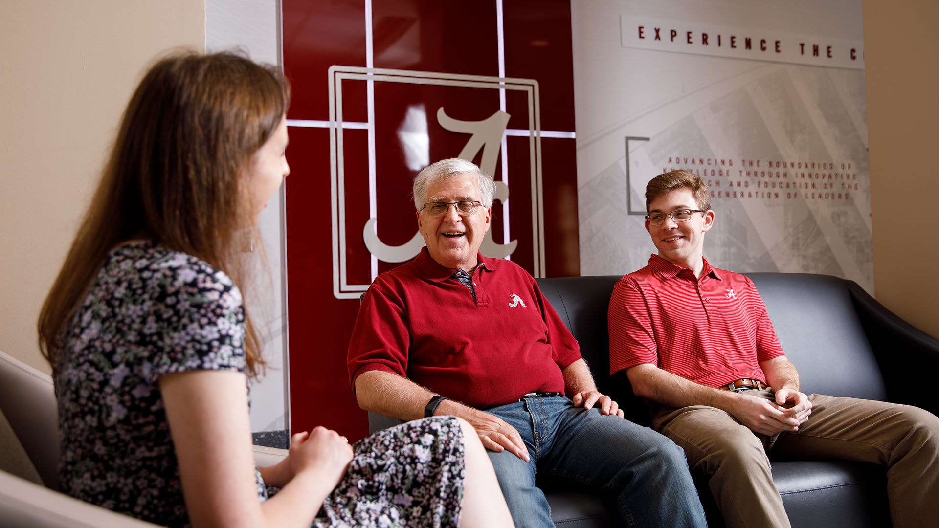 Bob Brazeal and 2 students sit chatting in a room with the UA logo on the back wall