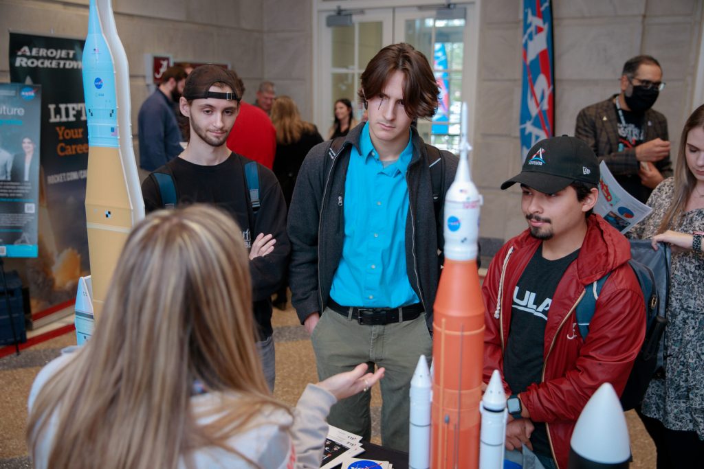 3 students listen to a booth presenter with a model rocket in the foreground