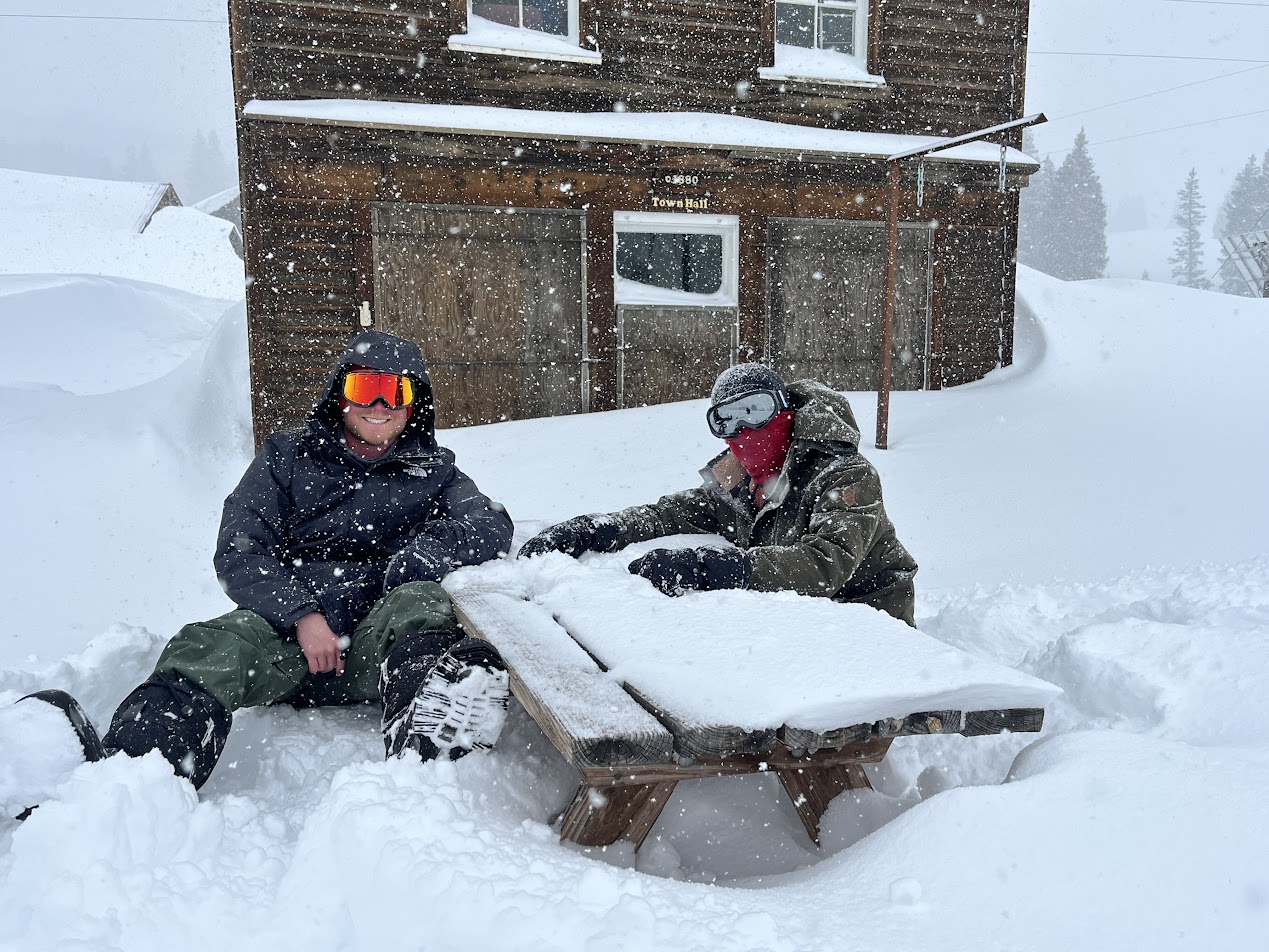 Two men sit at a snow covered picnic table.