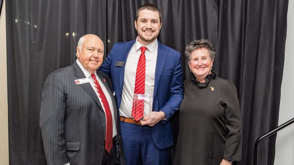 A white man in a blue suit and red tie holding his award in one hand with a man in a red tie and a woman in a black dress